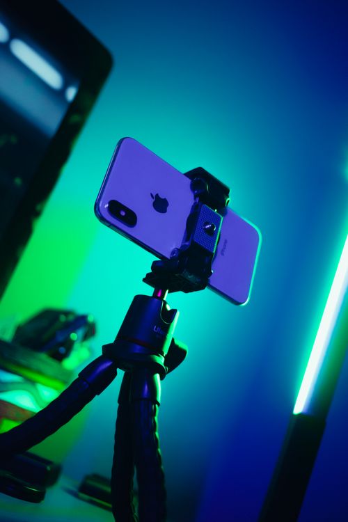 Free A smartphone mounted on a tripod, illuminated by neon blue and green lights. Stock Photo