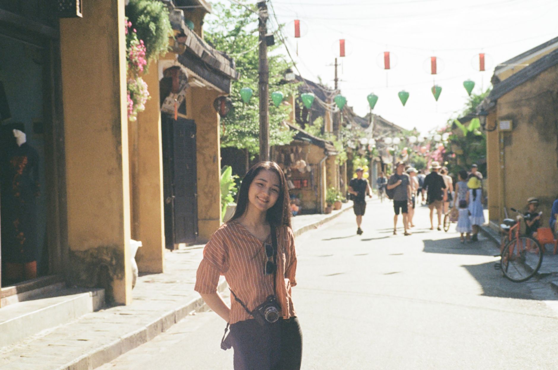 A woman with a camera smiles while exploring a sunny, vibrant street with lanterns and tourists in the background.