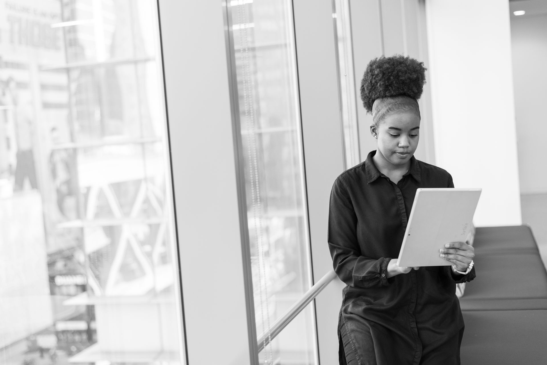 Black and white photo of a woman reading a tablet by a window in a modern workplace setting.