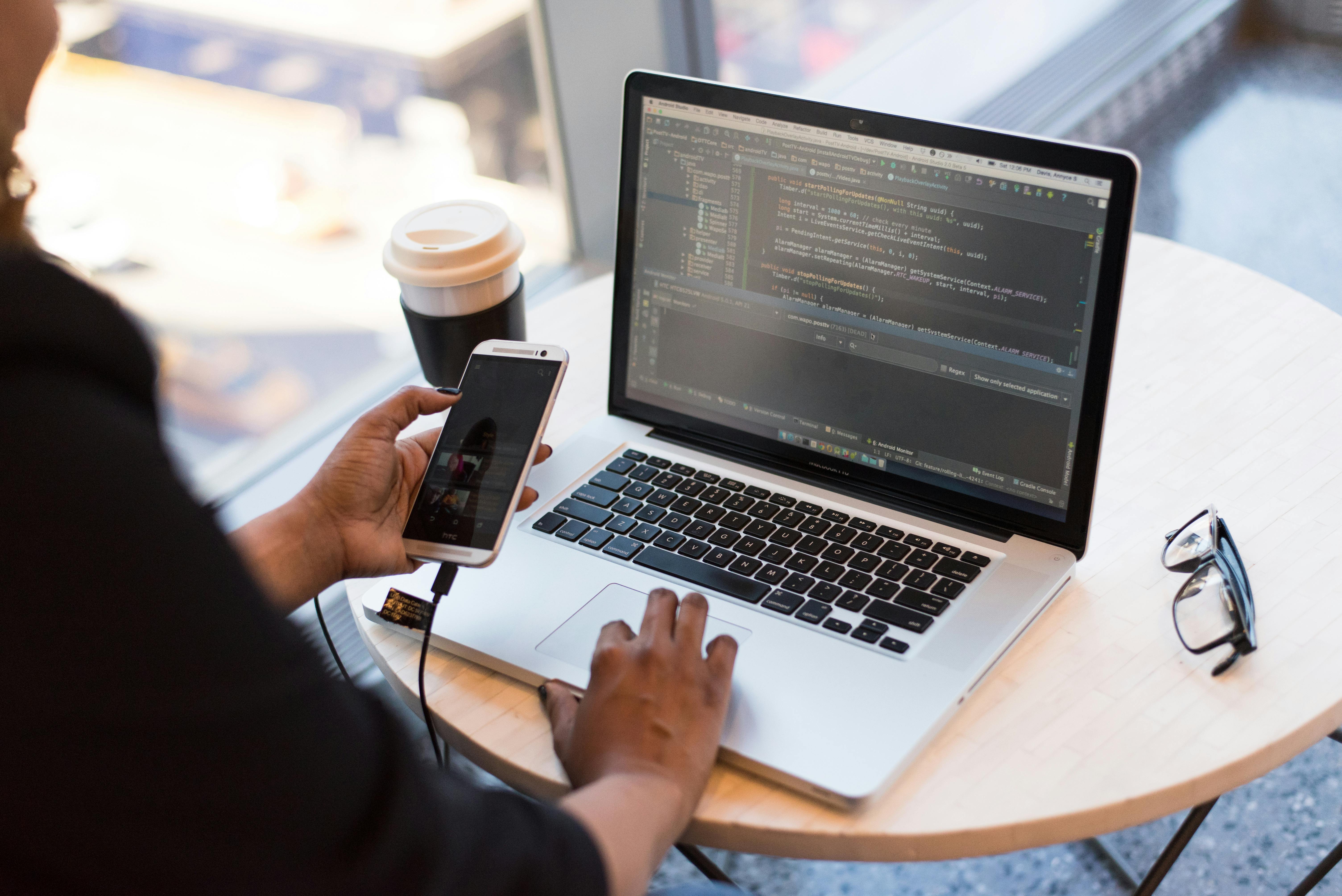 Woman looking at her mobile phone while using her laptop on the table. | Photo: Pexels