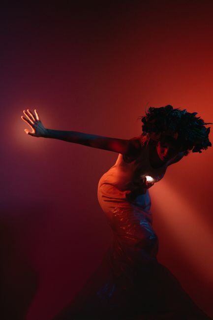 A woman in a floral crown bends gracefully under warm red lighting in a studio setting.