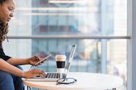 一位专注的黑人女性在明亮现代的办公场所使用笔记本电脑和手机，展现了多任务处理与远程工作的场景