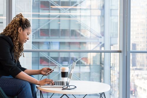 Free A woman working on a laptop with a smartphone, illustrating modern remote work. Indoor setting. Stock Photo