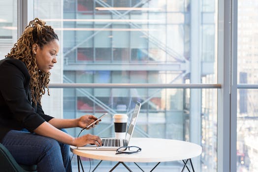 African American woman working remotely with a laptop and smartphone in a modern office setting.