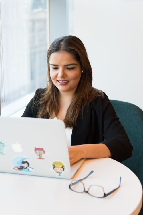 Free Smiling woman in black blazer working on laptop with open notebook on desk in a modern office setting. Stock Photo
