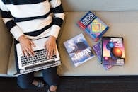 一个女人坐在沙发上，用笔记本电脑学习编码，周围堆满了编程书籍