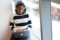 一位非裔美国妇女戴着耳机，在室内阳光明媚的窗前使用着笔记本电脑