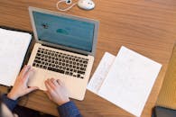 俯视视角下，一个人正坐在办公桌前使用笔记本电脑工作，桌面上散落着文件和纸张