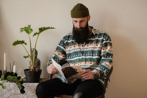 Free A bearded man in a patterned shirt reading a book, creating a relaxed and cozy atmosphere indoors. Stock Photo