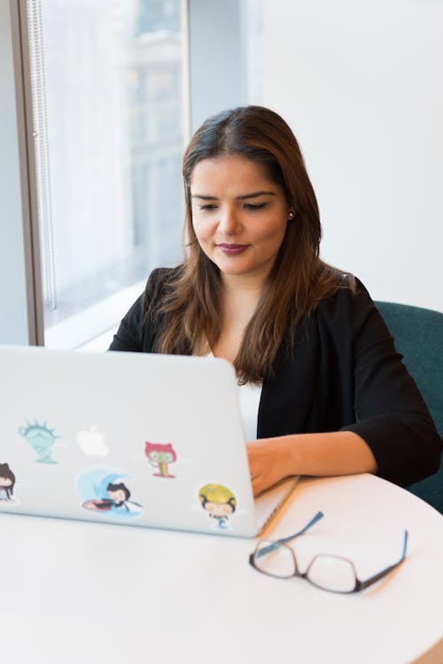 Woman Wearing Black Suit Jacket Using White Macbook With Stickers on White Wooden Table