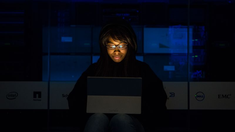 A woman uses her laptop in a dimly lit server room, focusing on technology and work.