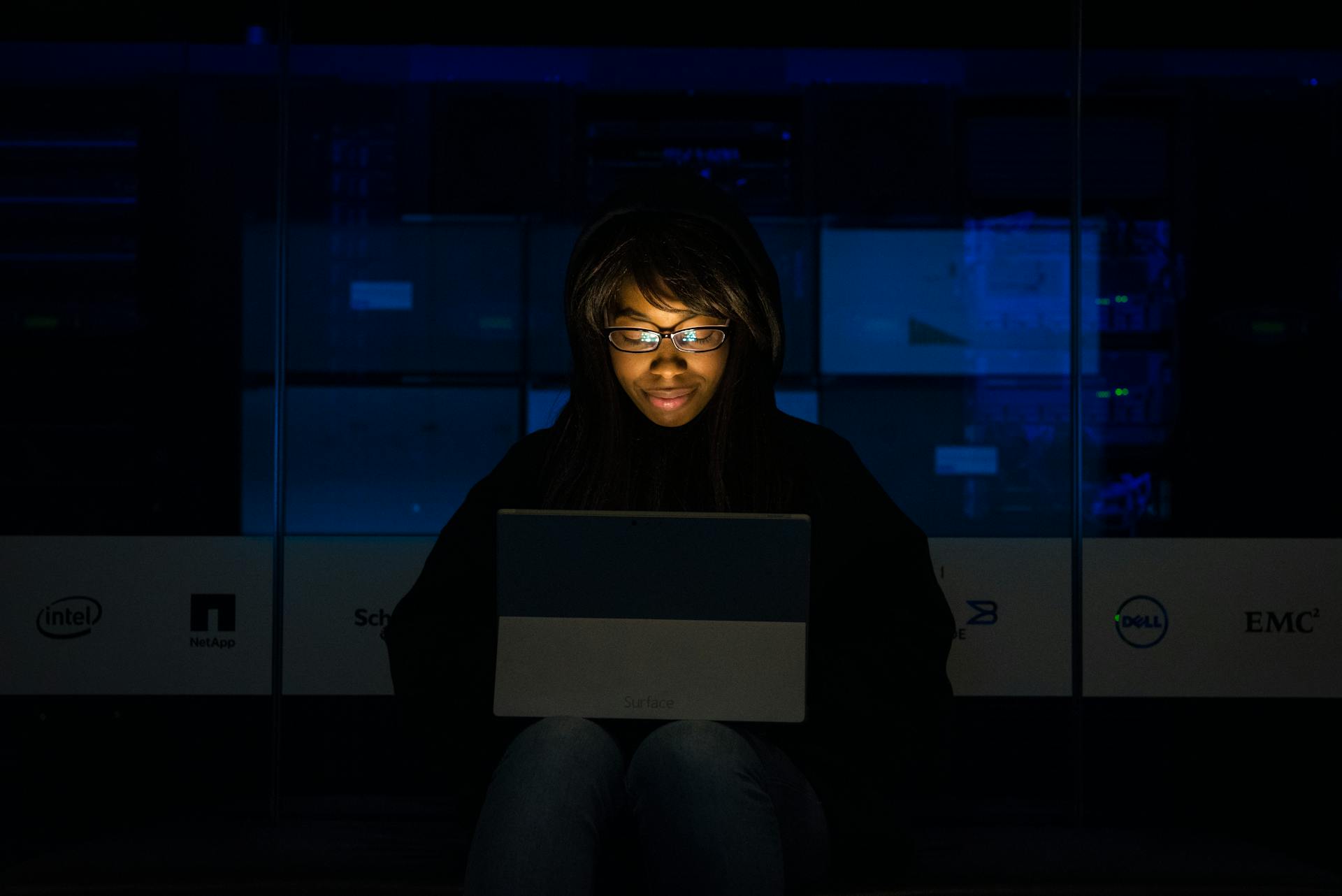 A woman uses her laptop in a dimly lit server room, focusing on technology and work.