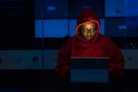 一名身穿红色连帽衫的女子在室内紧张地用笔记本电脑工作，屏幕的灯光照亮了她