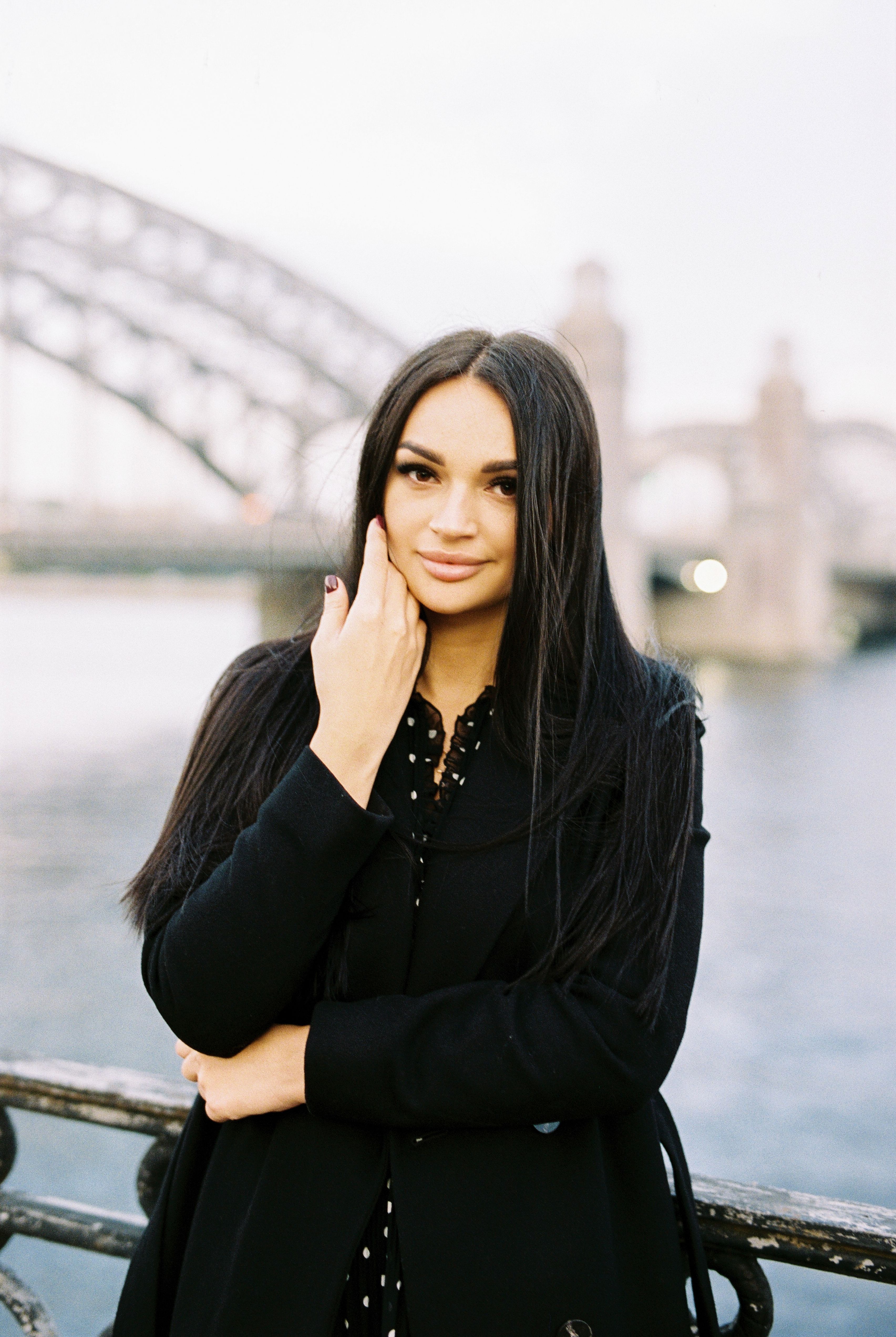 Elegant woman in black coat poses by riverside with iconic bridge in the background.