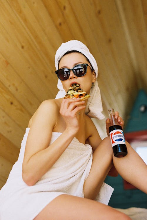 Woman in Towels and Sunglasses Holding Soda Bottle and Eating Pizza