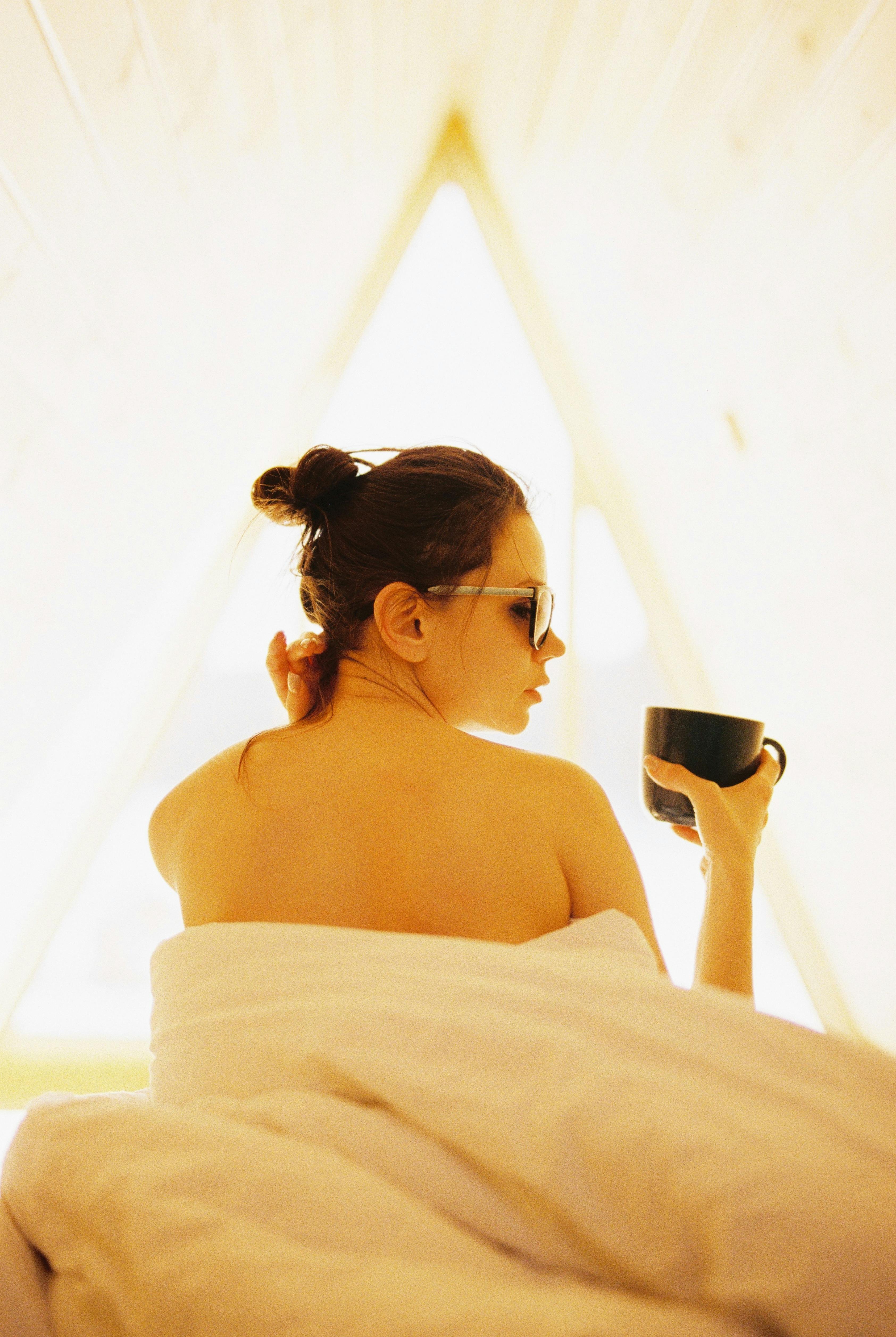 woman in sunglasses and tied up hair holding a mug while sitting in bed