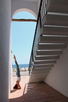 A woman practices a yoga headstand under a staircase by a sunny seaside.