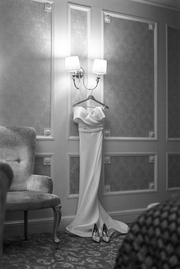 Wedding Dress On A Hanger Under The Lamps In A Hotel Room