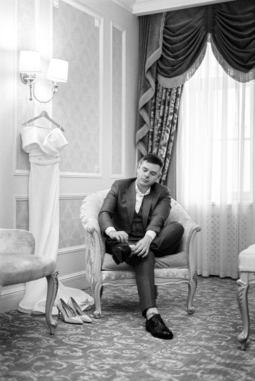 Free Young groom tying shoes in a luxurious suite, wedding attire nearby, black and white photo. Stock Photo