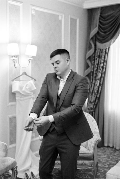 Free Portrait of a groom adjusting his sleeve in a classic hotel suite, suit and gown in view. Stock Photo