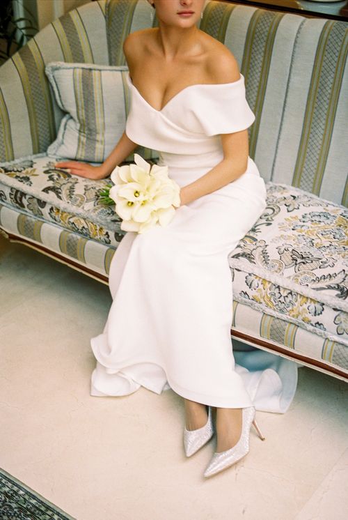 Free A beautiful bride in an off-the-shoulder gown, holding white flowers, sitting on a vintage sofa. Stock Photo