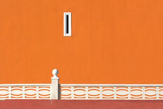 Minimalist orange wall with white patterned railing and a small window in urban setting.
