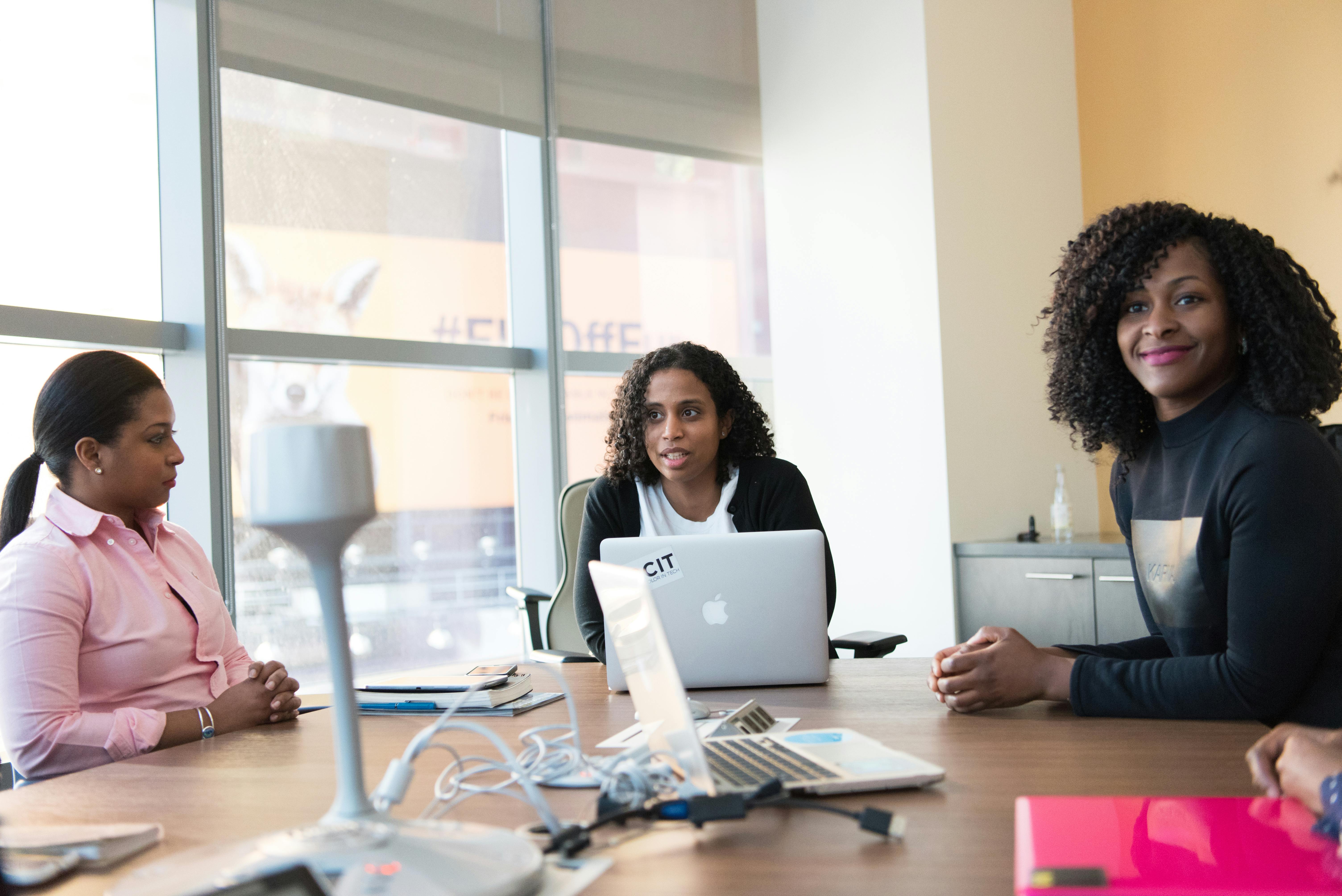 three woman sitting beside brown wooden conference table with silver apple macbook