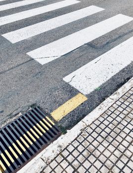 From above of asphalt road with uneven surface and cracks with similar wide lines representing pedestrian crossing near sewer manhole and dirty sidewalk in town