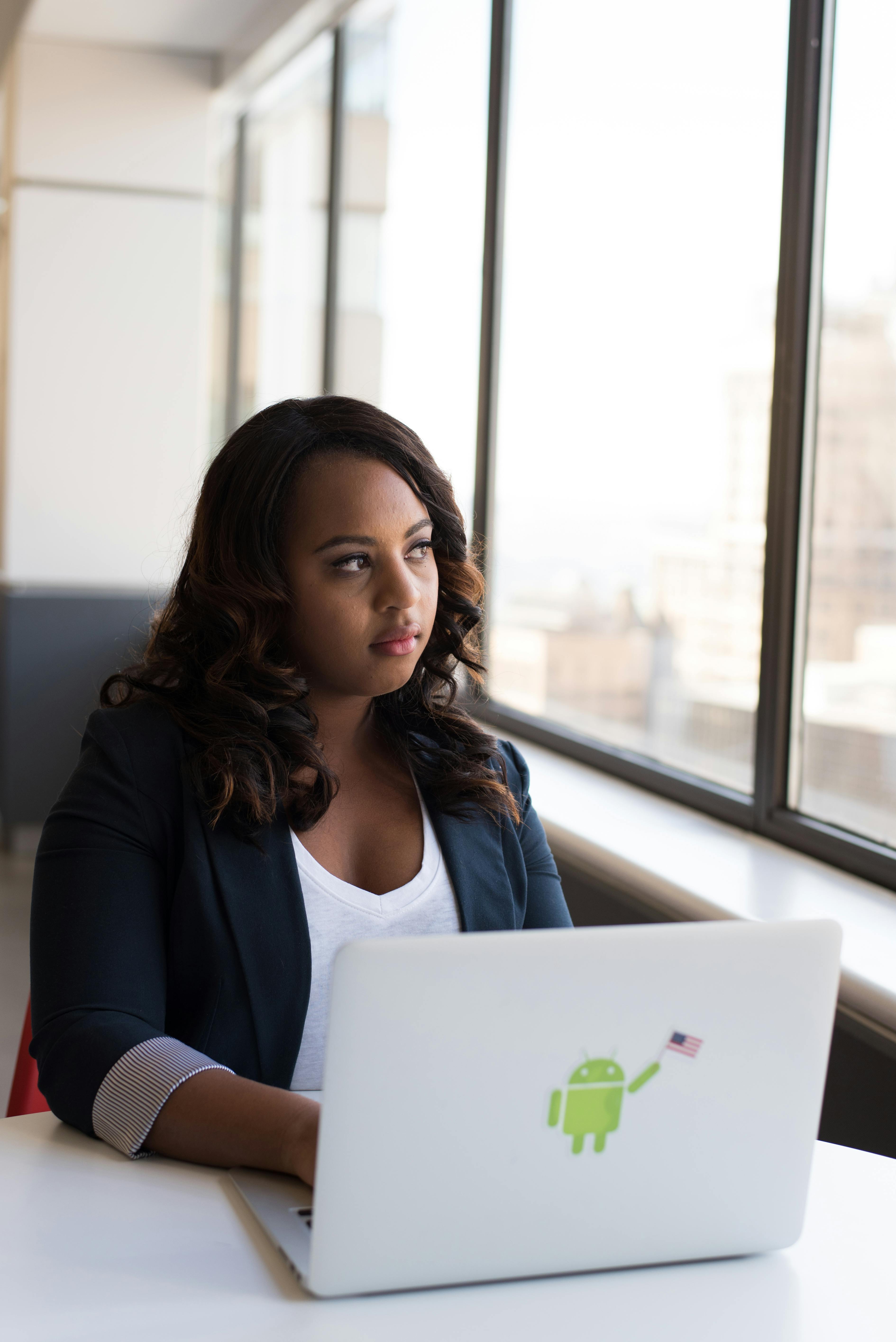 Focused woman using a laptop with Android sticker, sitting by a window in an office.