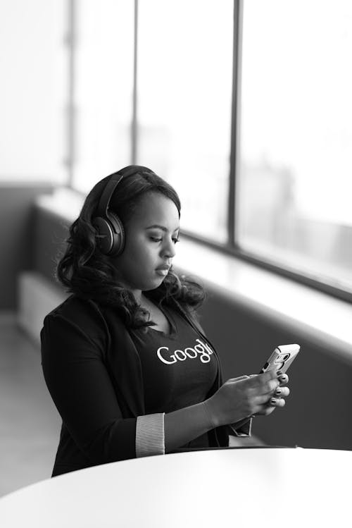 Grayscale Photo of Woman Holding Smartphone Wearing Wireless Over-ear Headphones