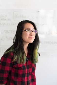 Portrait of a young woman with glasses standing indoors, wearing a red checkered shirt.
