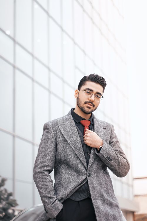 Free A confident man with eyeglasses poses in a stylish suit against a modern glass building. Stock Photo