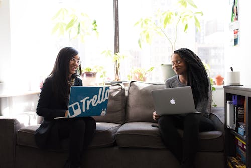 Two Women Sitting on Gray Couch Holding Laptop Computers