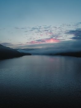 A serene lake reflecting a colorful morning sky during sunrise with misty clouds.