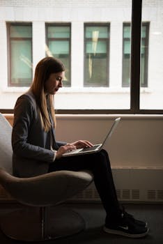 Woman sitting on a chair using a laptop by the window, engaged in remote work.