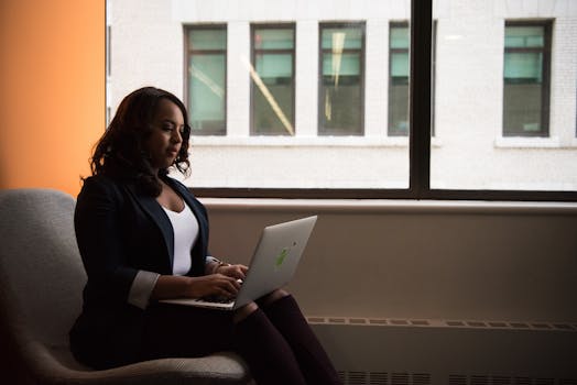 African American woman sits by window using a laptop, symbolizing modern remote work.