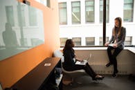 在现代办公环境中，两名妇女正参加商务会议，她们面前摆放着笔记本电脑，窗外是城市景观