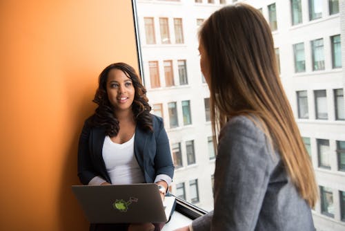 Free Woman Wearing Gray Blazer Facing Woman Wearing White Shirt and Black Blazer With Laptop on Lap Stock Photo