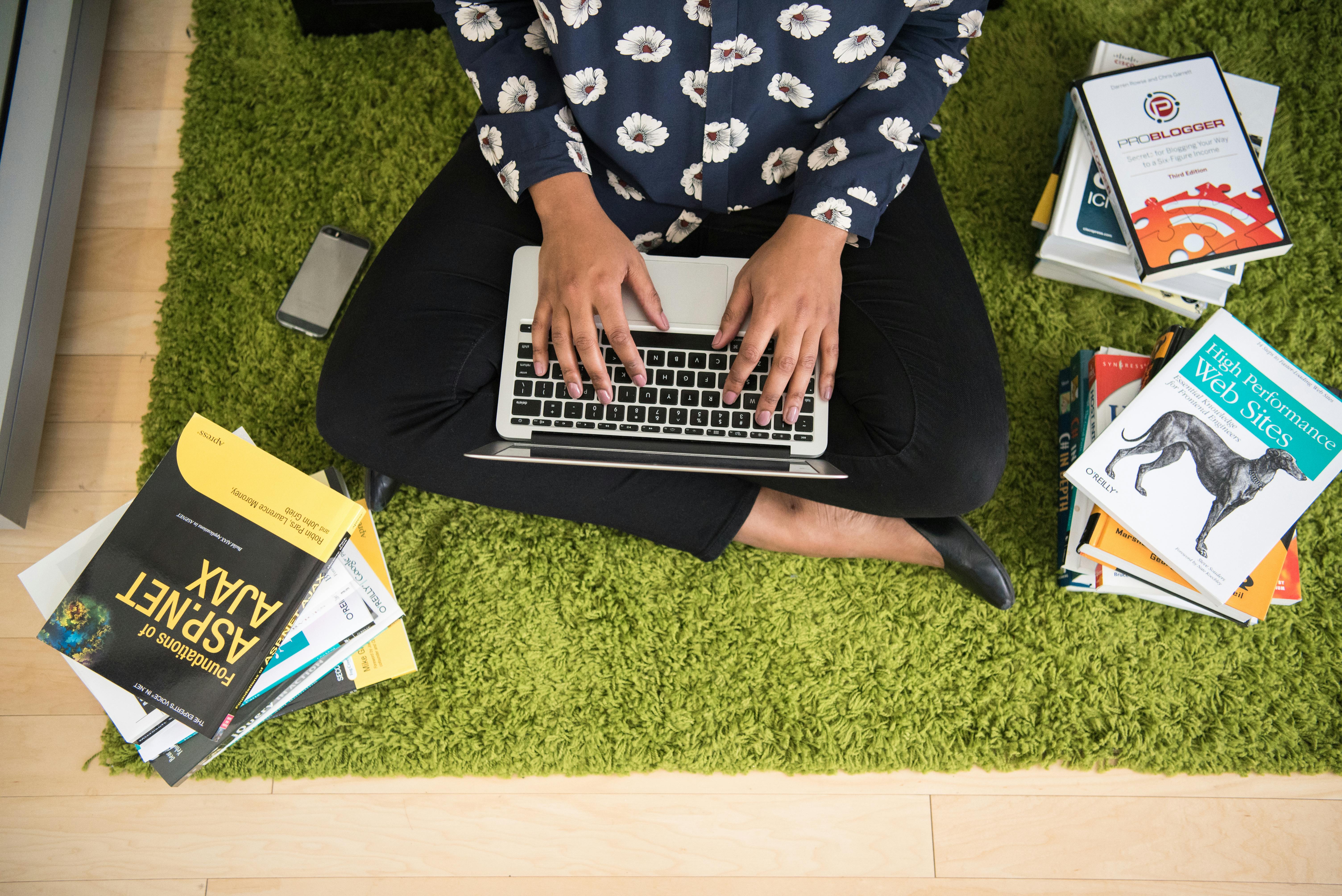 A woman sits on a green carpet working on a laptop with stacks of books around her, focusing on technology projects.