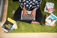 一名妇女坐在绿色地毯上，专注地在笔记本电脑上处理技术项目，周围堆满了书籍