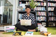 一名妇女坐在图书馆的地毯上，用笔记本电脑工作，周围堆满了书