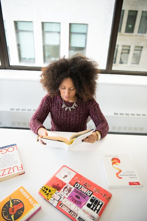 Free Woman in Maroon Sweatshirt Reading Book on White Wooden Table Near Clear Glass Window Stock Photo