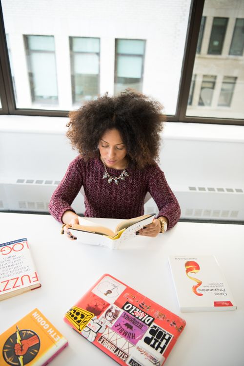 Free A woman engaged in reading a book at a white desk, surrounded by colorful books, in a modern office space. Stock Photo