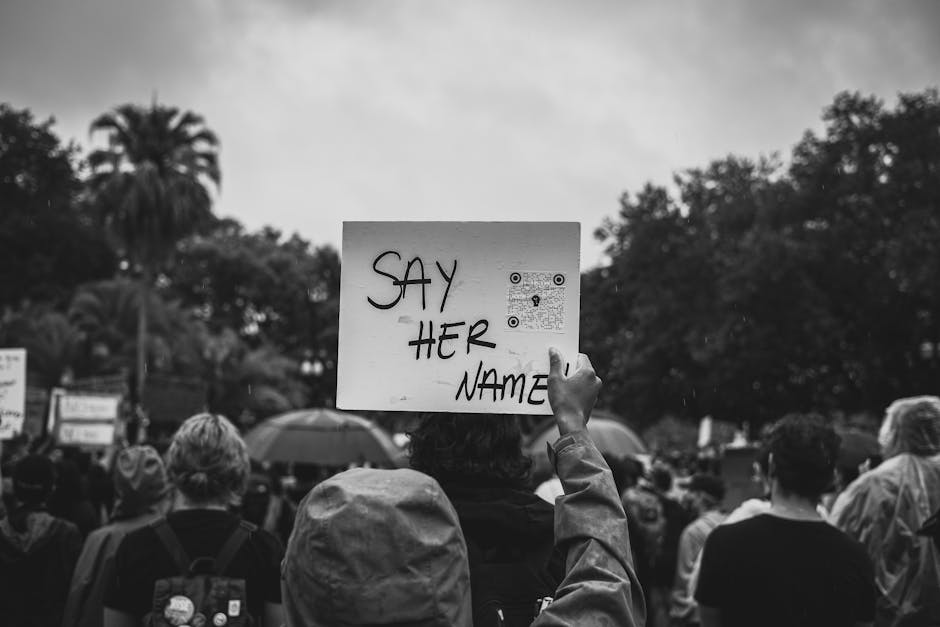 Black and white image of a protest with a 'Say Her Name' sign raised in a crowd.