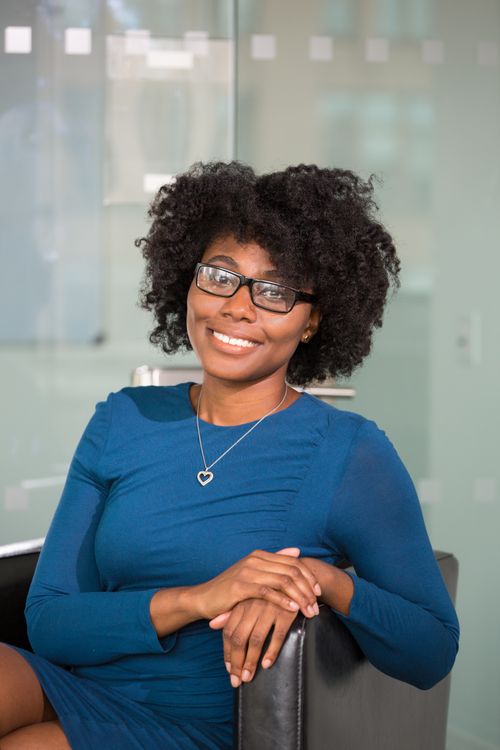 Free Portrait of a smiling woman with curly hair and glasses in a blue dress sitting indoors. Stock Photo