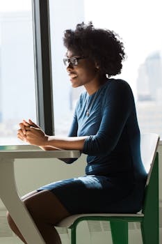 A joyful African American woman with glasses sitting indoors by a window, enjoying daylight.
