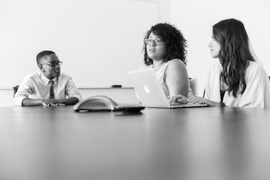 Three professionals discussing work at a conference room table with laptops in a modern office.