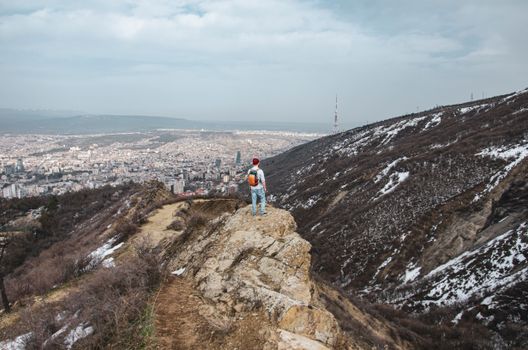 A traveler stands on a snowy mountain edge with a panoramic view of Tbilisi cityscape.