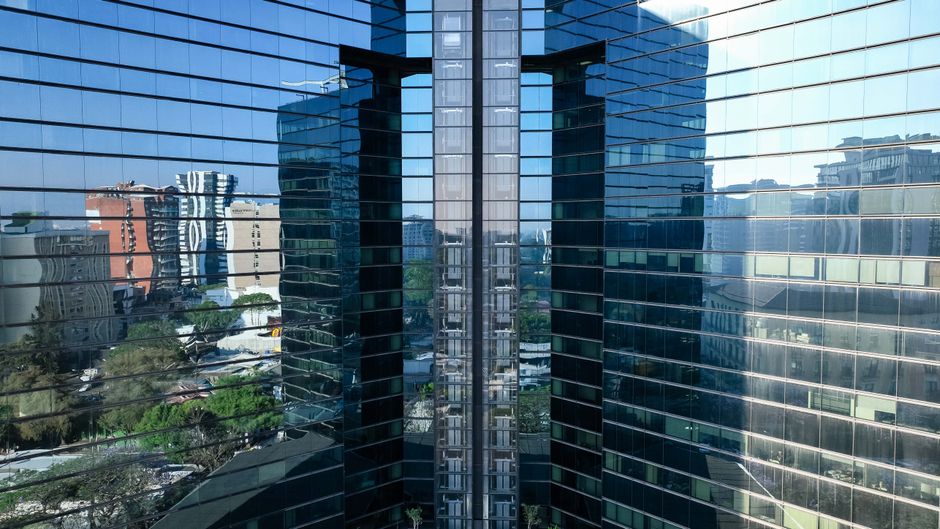Fotomural Edificio alto revestido de cristal reflejando el horizonte urbano a plena luz del día.