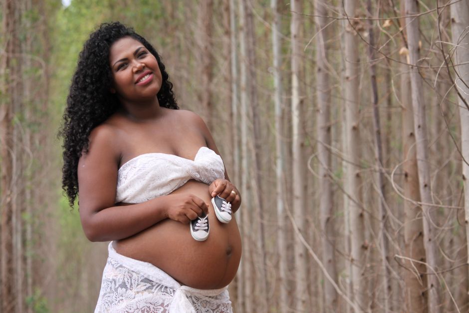 5 Maneiras de Cultivar a Amizade Feminina na Maternidade 2 Smiling pregnant woman holding baby shoes in a serene forest setting.